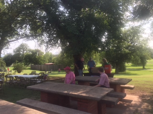 A group of people standing around a picnic table in a park.